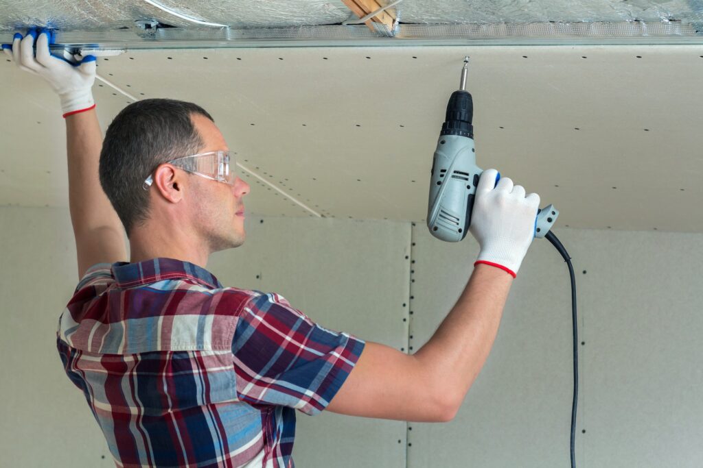 Young man in goggles fixing drywall suspended ceiling to metal frame using electrical screwdriver on